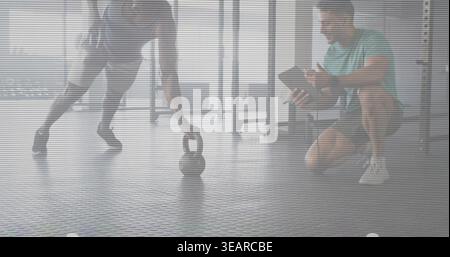 Longierender Auszubildender, der die Kettlebell auf Gummiboden durch den Windows-Trainer in blauem Hemd mit Tablette erreicht. Fitnessstudio, Training, Coaching, Gewichte, Fitness, studio Stockfoto