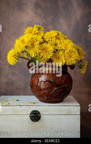 Vertikales Stillleben von gelben Löwenzahn in einem rustikalen Tontopf, der auf einer alten weißen Holzkiste liegt. Elegante Blumenkomposition in warmen Tönen. Stockfoto