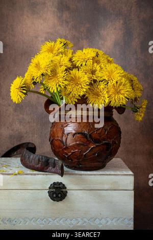 Vertikales Stillleben von gelben Löwenzahn in einem rustikalen Tontopf, der auf einer alten weißen Holzkiste liegt. Elegante Blumenkomposition in warmen Tönen. Stockfoto