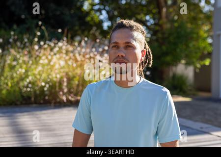 Afroamerikaner, der auf Holzdeck steht und hellblaues T-Stück trägt und auf hohe Gräser blickt. Dreadlocks, Porträt, Sonnenlicht, draußen, Promenade, Natur Stockfoto