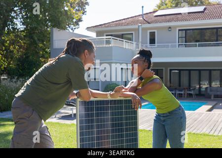 Ein Paar lehnt sich auf ein freistehendes Solarmodul im Hinterhof und plaudert am Pool und an der Terrasse. Sonnenlicht, Rasen, Terrasse, Dachterrasse, erneuerbar, Energie, modern Stockfoto