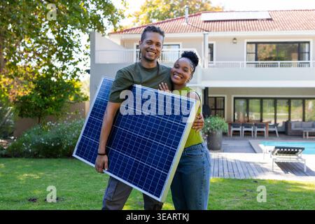 Afroamerikanisches Paar, das auf dem Rasen vor dem Haus steht und Solarzellen hält, lächelnd. Photovoltaik, Energie, erneuerbar, nachhaltig, Wohnraum, außen Stockfoto