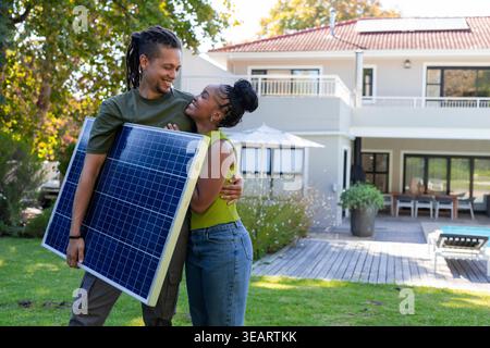 Afroamerikanisches Paar hält einen großen Solarpanel und umarmt sich auf dem Rasen vor dem modernen Haus. Photovoltaik, im Freien, sonnig, Terrasse, Hof, Öko Stockfoto