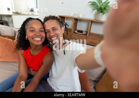 Afroamerikanisches Paar lächelt und nimmt Selfie, während es zu Hause auf der Couch mit Smartphone sitzt. Sofa, Kissen, Pflanze, Regale, Dekor, modern, gemütlich Stockfoto