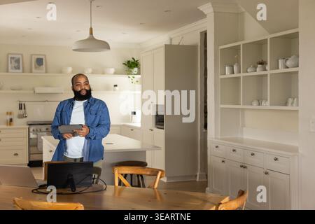Erwachsener afroamerikanischer Mann, der Denim-Hemd von der Kücheninsel trägt, der ein Tablet hält, das auf Laptop arbeitet. HomeOffice, Fernbedienung, modern, neutral, openplan, dini Stockfoto
