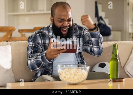 Afroamerikaner sitzt zu Hause auf der Couch und hält ein rotes Telefon, jubelt mit Popcorn und Bier. Smartphone, Soccerball, gemütlich, neutral, lässig, offen Stockfoto