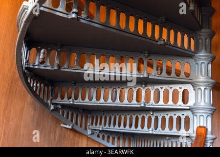 Nahaufnahme einer kunstvollen Gusseisenspiraltreppe vor warmer Holzvertäfelung, die geschwungene Stufen, dekorative Baluster und Industrie hervorhebt Stockfoto