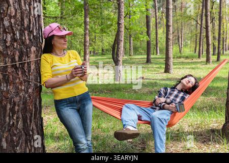 Teenager, der in der Hängematte schläft, während Mutter im Wald Kaffee genießt Stockfoto