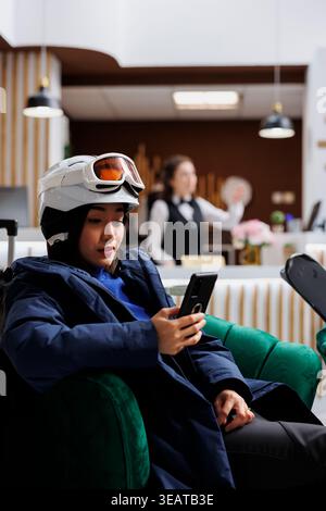 Während der Winterferien benutzt die junge asiatische Frau das Handy in der gemütlichen Lounge. Weibliche Gäste, die auf einem Sofa mit digitalem Smart-Gerät sitzen, und Skiski mit Winterjacke Skibrille und Helm tragen. Stockfoto