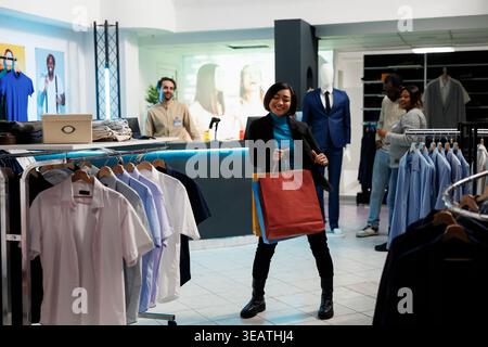 Lächelnde asiatische Frau tanzt mit Einkäufen und genießt es, Kleidung im Bekleidungsgeschäft zu kaufen. Fröhliche Boutique-Gäste, die Pakete halten und im Einkaufszentrum Musik spielen Stockfoto