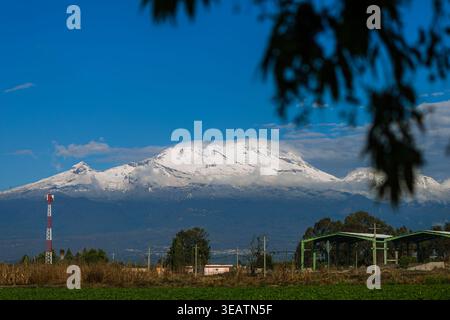 Majestätischer Panoramablick auf den schneebedeckten Berg Iztaccihuatl mit grünen Feldern und einem Kommunikationsturm im Vordergrund. Stockfoto
