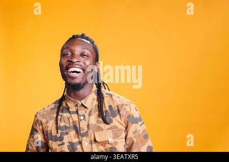 Porträt eines afroamerikanischen Mannes, der während des Fotoshootings lächelt und vor der Kamera im Studio auf gelbem Hintergrund lacht. Entspannte, fröhliche Person mit positivem Gesichtsausdruck, posiert selbstbewusst Stockfoto