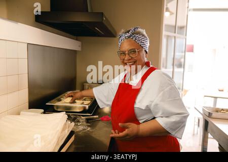 Glückliche Seniorin in roter Schürze, die in der Küche steht, während sie Essen zubereitete Stockfoto