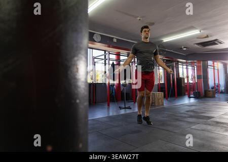 Mittelgroßer Mann, der im Fitnessstudio mit Schlafsack, Pull-Up-Rigs, Plyboxen und Tageslichtfenstern springt Stockfoto