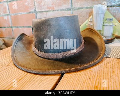 Valmiera, Lettland - 22. April 2026: Stilvoller Cowboyhut aus Leder auf einem Holztisch in einem gemütlichen rustikalen Café. Warme Töne und strukturierte Backgr Stockfoto