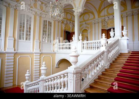 Elegantes Interieur des Rundāle-Palastes in Lettland mit einer kunstvollen barocken Treppe mit weißen Brüstungen, dekorativen Säulen und einem roten Teppich. Stockfoto