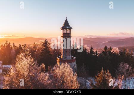 Der Stepanka-Aussichtsturm erhebt sich über die Winterlandschaft im Isergebirge. Sonnenuntergänge erzeugen warmes Licht über Bäumen und Hügeln. Besucher genießen die malerische Aussicht vom Turm bei Sonnenuntergang. Stockfoto