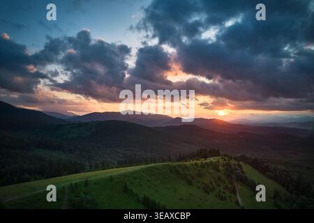 Atemberaubender Sonnenuntergang über dem Wald aus der Vogelperspektive. Sonnenstrahlen brechen durch dramatische Wolken und strahlen goldenes Licht über sanfte Hügel und Täler. Ferne Berge schaffen eine ruhige und malerische Szene. Stockfoto