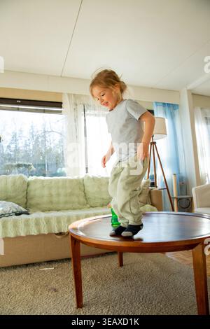 Niedrigwinkelaufnahme eines fröhlichen kleinen Kindes mit langen Haaren, das vom Sofa springt, voller Energie und verspieltem Lachen während eines hellen morgens zu Hause Stockfoto