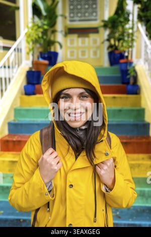 Glückliche junge Frau, die einen leuchtend gelben Regenmantel und eine Mütze trägt, lächelt in die Kamera und steht vor leuchtenden, mit Regenbogen bemalten Außentreppen in A c Stockfoto