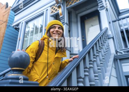 Frau mit gelbem Regenmantel und Beanie lächelnd, während sie die Außentreppe eines klassischen viktorianischen Hauses in san francisco hinaufsteigt und die Essenszeit festnimmt Stockfoto