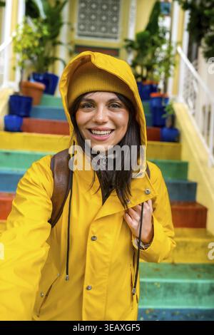 Frau lächelt und genießt Städtereise, trägt eine leuchtende gelbe Regenjacke und Mütze, während sie auf einer farbenfrohen Regenbogentreppe in san steht Stockfoto
