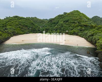 Blick aus der Vogelperspektive auf den abgeschiedenen Sedahan Beach, eine sandige Bucht umgeben von grünen Hügeln und stürzenden türkisfarbenen Wellen, Gunungkidul Regency, Indonesien. Stockfoto