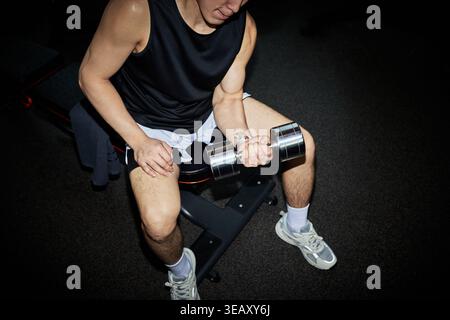 Hochwinkelansicht eines jungen, erwachsenen asiatischen Mannes, der sitzende Hantelcurl im Fitnessstudio durchführt und Krafttraining und Fitness-Routine zeigt. Ideal für Trainingsaktionen, Wellness-Marketing, Sportanzeigen Stockfoto