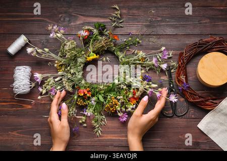 Frau mit Vorräten, die einen Kranz aus schönen Wildblumen auf braunem hölzernem Hintergrund herstellt. Sommersonnenwende Stockfoto