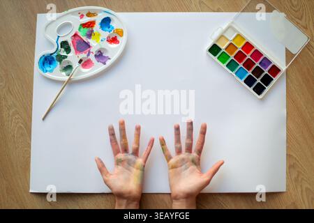 Kinder, Die Hände Mit Leerem Papier Malen. Blick von oben auf bemalte Kinderhände, leeres Papier, Aquarellset, Pinsel und Palette für kreative Kunst Stockfoto