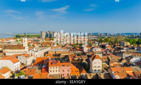 Blick aus der Vogelperspektive auf das historische Viertel Zemun mit rot gekachelten Dächern, den St. Nikolaus Kirchturm und die moderne Skyline der Stadt unter einem klaren Blau Stockfoto