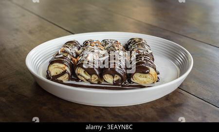 Nahaufnahme von köstlichen Crepes mit Schokoladensoße und mit Haselnüssen bestreut auf weißem Teller auf rustikalem Holzbrett. Dünne Dessertcrepes, RO Stockfoto