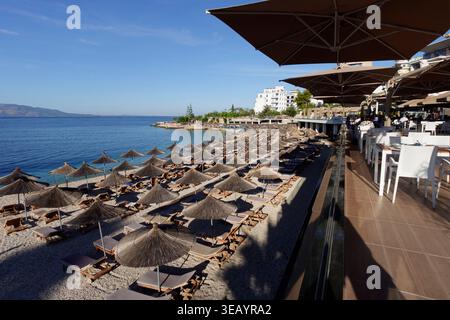 Strohschirme und Liegestühle an einem Strand neben einer Restaurantterrasse am Wasser in einem Sommerresort in Sarandë, Albanien Stockfoto