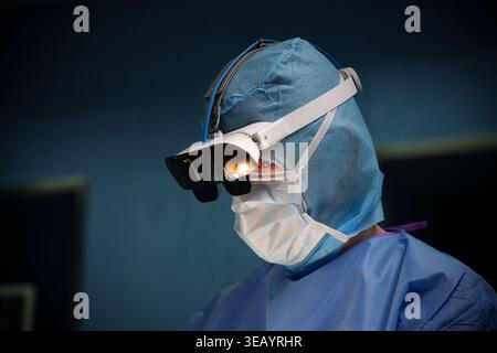 Männlicher Chirurg in blauen sterilen Peelings mit Operationslupen und Stirnlampe während eines ärztlichen Eingriffs Stockfoto