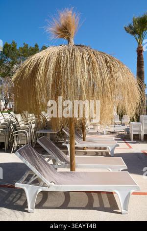Reihe leerer weißer Sonnenliegen unter traditionellen Strohschirmen in einem mediterranen Resort-Hotel mit Palmen unter klarem blauem Himmel im Sommer Stockfoto