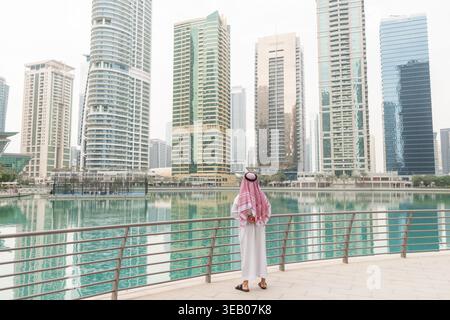 Ein arabischer Geschäftsmann steht tagsüber am Wasser und betrachtet hohe Gebäude in einer Stadt im Nahen Osten. Er trägt traditionelle Kleidung. Stockfoto