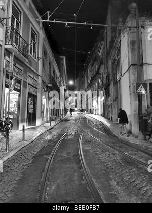 Nächtlicher Blick auf eine Gasse in der Altstadt von Lissabon, gepflasterte Gasse mit Straßenbahnschienen, umgeben von beleuchteten historischen Gebäuden in tiefem Schwarz-weiß Stockfoto