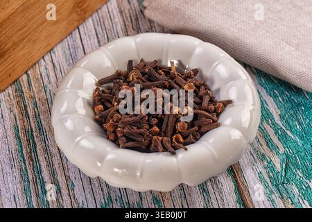 Eine Nahaufnahme ganzer getrockneter Nelken in einer weißen Keramikschale. Das aromatische Gewürz wird auf einer rustikalen, mehrfarbigen Holzoberfläche mit einem Leinen präsentiert Stockfoto