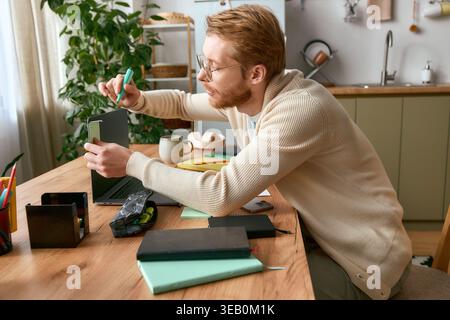 Der junge Mann in einem leichten Pullover konzentriert sich auf das Basteln von Ideen an seinem einladenden Schreibtisch im Home Office. Stockfoto