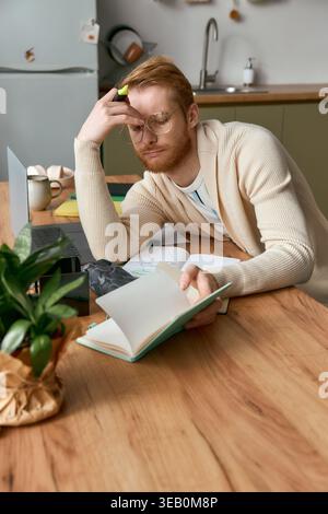 Ein junger Mann mit Brille reflektiert besinnlich, während er in einer schicken Küche ein Buch liest. Stockfoto