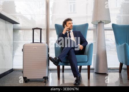Geschäftsmann, der im Lobbystuhl des Hotels sitzt und neben dem Gepäck auf dem Handy spricht Stockfoto