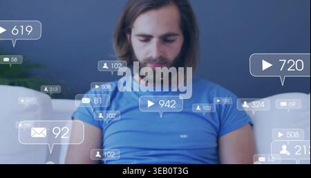Sitzender bärtiger Mann in blauem T-Stück, der auf weißem Sofa auf den Schoß blickt, mit Sozialstatistiken, Kissen, Pflanze Stockfoto