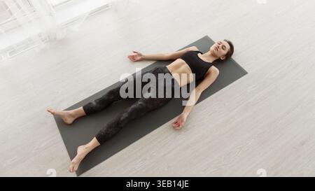 Frau, die tagsüber Entspannung übt, posiert auf der Yoga-Matte in einem hellen Innenraum Stockfoto