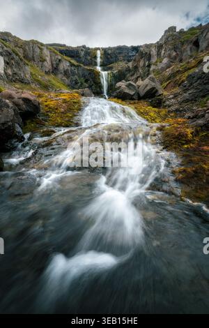 Blick auf einen kaskadierenden Wasserfall, der über moosige Felsen und zerklüftete Klippen unter einem bewölkten Himmel fließt Eskifjordur, Fjardabyggd, Island. Stockfoto