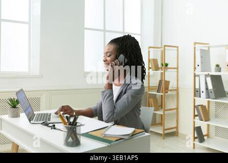 Erfolgreiche Geschäftsfrau, die ihr Telefon hält, während sie in modernen Büroumgebungen an einem Laptop arbeitet Stockfoto