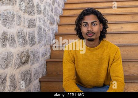 Indischer Mann sitzt auf einer Holztreppe in der Nähe einer strukturierten Steinmauer, trägt Senfpullover und Jeans Stockfoto