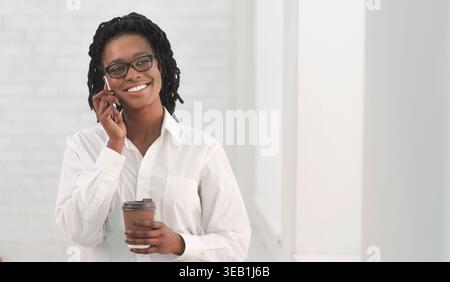 Lächelnde Frau am Telefon hält Kaffee in modernem hellen Raum Stockfoto