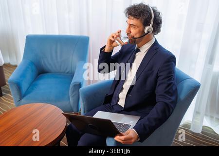 Business Professional Trinkwasser während der Arbeit an einem Laptop mit Headset in der komfortablen Hotel Lounge Stockfoto