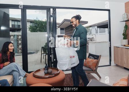 Mann, Der Eine Einkaufstasche An Eine Frau Im Wohnbereich Übergibt, Wiederverwendbare Einkaufstasche Und Lächelnde Gesichter, Offene Glastüren Zur Terrasse Und Gefliestem Boden, Leder Stockfoto