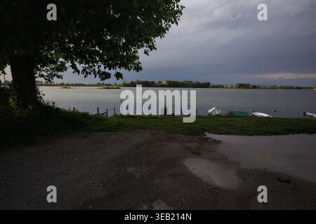 Lakeshore mit verankerten Booten, von etwas über einem Pier aus gesehen, mit Pfützen auf dem Boden, in der Dämmerung vor einem Sturm, eingerahmt von einer Pappel Stockfoto